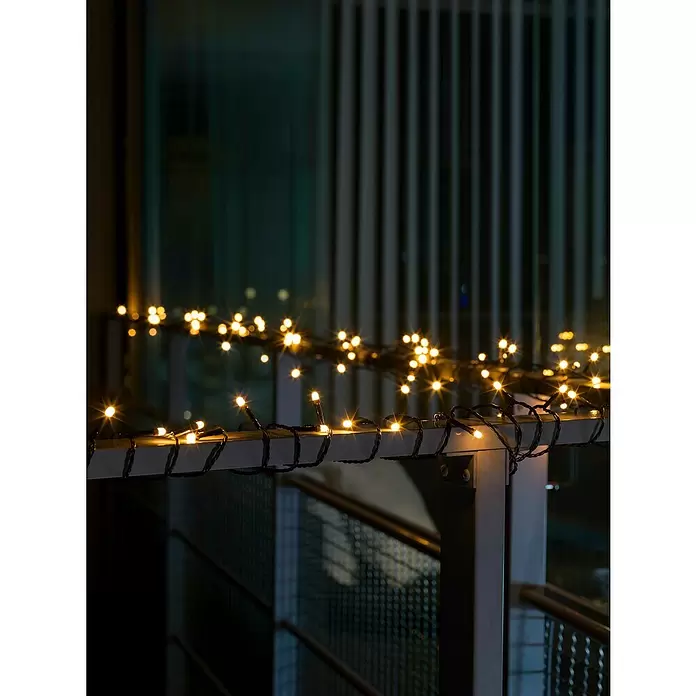 Konstsmide LED-Lichterkette - Lichtfarbe: Bernstein, Anzahl Leuchtmittel: 120 Stk., Netzbetrieben, IP44 - Warmweiße Lichterkette mit dunklen Kabeln, nachts um ein Balkongeländer gewickelt, erzeugt ein gemütliches Licht.