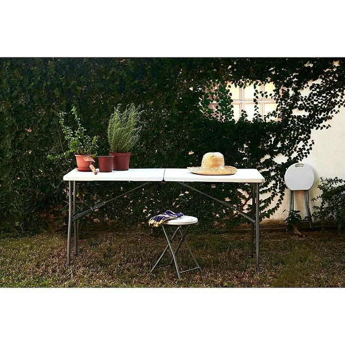 Mesa de jardín Every - L x An x Al: 152 x 70 x 74 cm, Acero/HDPE, Blanco roto, Abatible - Escena de jardín con una mesa plegable robusta, perfecta para trasplantar, complementada con plantas, un sombrero y un taburete plegable.