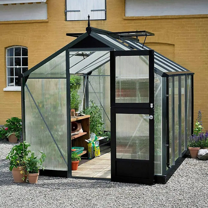 Juliana Gewächshaus Compact - Außenmaß inkl. Dachüberstand (B x T): 224 x 295 cm, Material Seitenwand: Kunststoff, 10 mm, Anthrazit/Schwarz - Ein geräumiges Gewächshaus aus Polycarbonat mit einer offenen Tür, umgeben von Pflanzen und Kies.