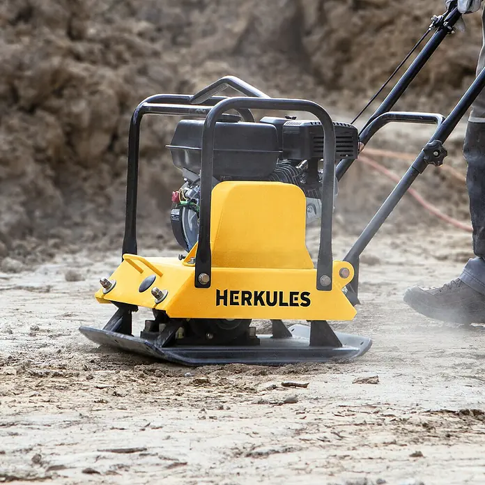 Herkules Rüttelplatte RP1800 - 4,8 kW, Verdichtungsdruck: 2.300 kg - Bild einer gelben Herkules-Rüttelplatte, die auf einem staubigen Untergrund steht und von einer Person bedient wird.