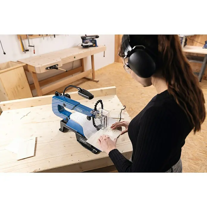 Scheppach Dekupiersäge SD 1600 V - 120 W - Eine Frau arbeitet mit einer Scheppach Dekupiersäge an einem Holzmuster in einer Werkstatt, wobei sie Kopfhörer und Schutzbrille trägt.