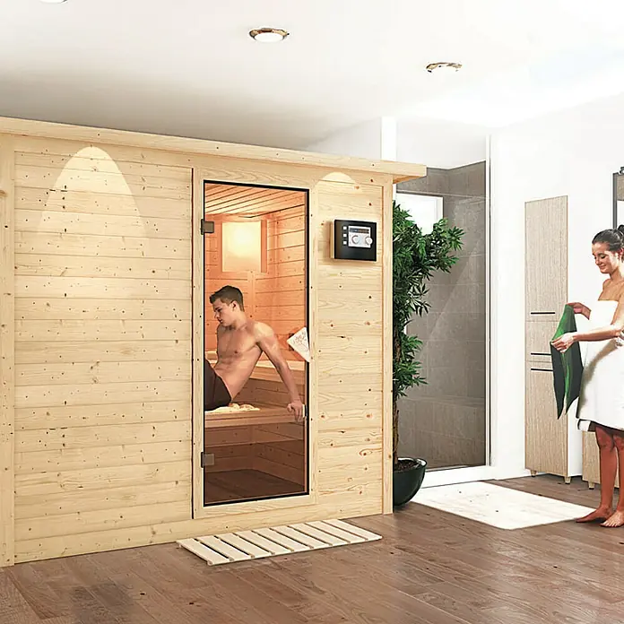 Karibu Massivholzsauna Sonara - Mit 9 kW Bio-Ofen mit externer Steuerung, Mit Dachkranz und Beleuchtung, L x B x H: 259 x 210 x 192 cm - Entspannen Sie sich in dieser stilvollen Sauna. Ein Mann genießt die Wärme, während eine Frau sich mit einem Handtuch abtrocknet.