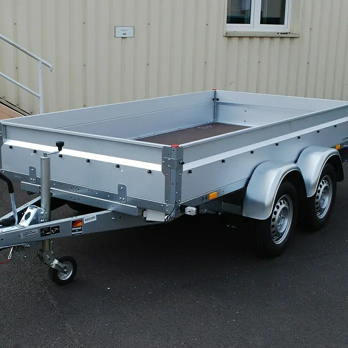 Stema Anhänger BASIC ST 2000-30-15.2 - Nutzlast: 1.575 kg, Doppelachser, Kasteninnenmaß: 301 x 153 x 35 cm, Gebremst - Silberner Pkw-Anhänger mit schwarzem Boden auf Asphalt vor einem Gebäude. Robuste Bauweise für vielfältige Transportaufgaben.