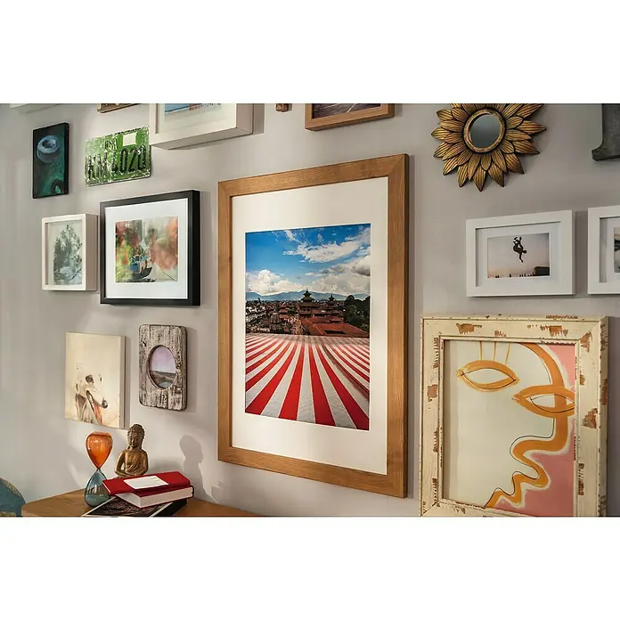 Tesa Selbstklebestrip CLICK COMBO 6X2 M & 8X2 L - 14 Stk., B x L: 20 x 85 mm - Die Wand ist mit einer Vielzahl von gerahmten Bildern, einer Spiegel-Sonnenblume und einer Lizenzplatte geschmückt. Die Rahmen variieren in Farbe und Stil und verleihen einen eklektischen Touch.