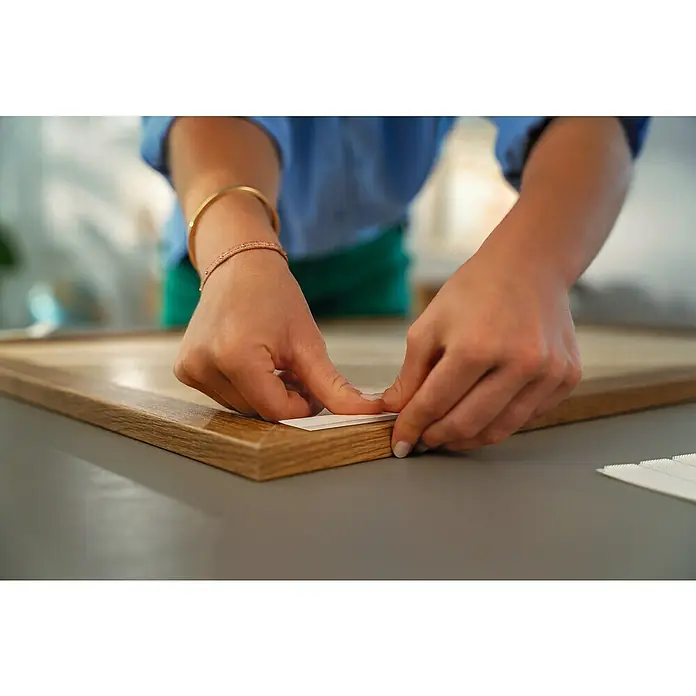 Tesa Selbstklebestrip POWERSTRIPS CLICK 4X2 S - 4 Stk., B x L: 20 x 60 mm - Nahaufnahme einer Person, die ein Stück Papier in einen Holzrahmen einfügt, vermutlich um ein Bild zu befestigen.