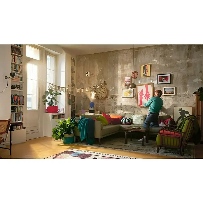 Tesa Powerstrips Selbstklebehaken - Eckig, Größe: S, Weiß, 3 Stk., Metall/Kunststoff - Ein gemütliches Wohnzimmer mit einer grauen Couch, bunten Kissen und einer "Happy Birthday"-Girlande an der Wand. Ein Mann hängt ein Bild auf.