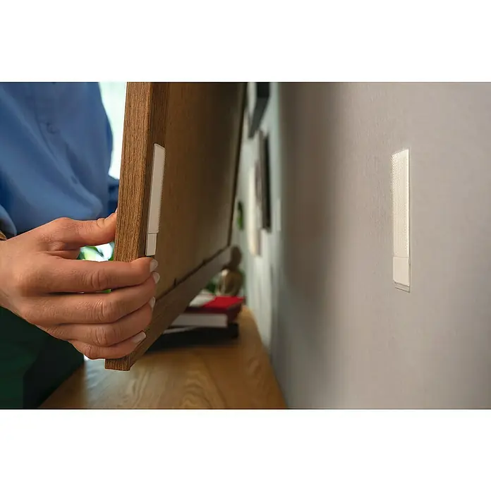 Tesa Selbstklebestrip CLICK COMBO 6X2 M & 8X2 L - 14 Stk., B x L: 20 x 85 mm - Nahaufnahme von einer Person, die mit Klebestreifen einen Holzrahmen an einer Wand befestigt.