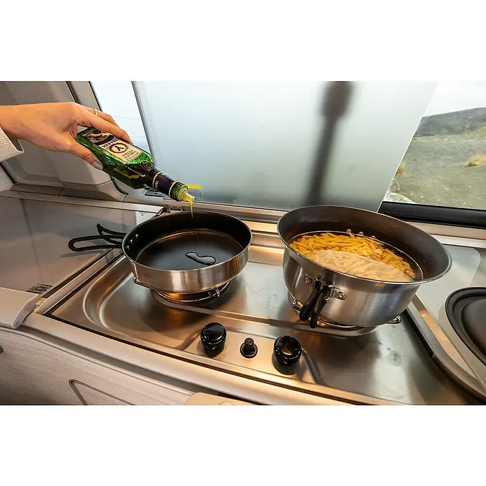 Petromax Kochgeschirr-Set Dimego - Edelstahl - Eine Hand gießt Olivenöl aus einer grünen Flasche in eine Bratpfanne auf einem Herd, neben einem Topf mit kochender Pasta.