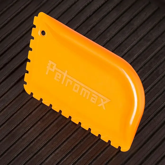 Petromax Schaber für Gusseisentöpfe - Kunststoff - Petromax Schaber: Ein orangefarbener Petromax Schaber zum Reinigen von Grillrosten, liegt auf einem schwarzen Grill. Das Werkzeug hat Zacken für eine effektive Reinigung.