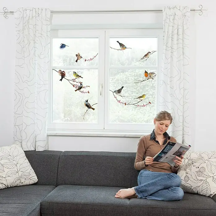 Komar Fenstersticker Birds - Bunt, 31 x 31 cm - Eine Frau sitzt auf einem Sofa und liest eine Zeitschrift vor einem Fenster, das mit dekorativen Vogelaufklebern versehen ist.