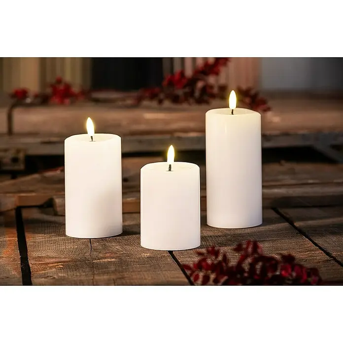Flower Power Vela con LED Novita - Blanco, Ø x Al: 7,5 x 10 cm, Cera natural - Primer plano de tres velas LED inalámbricas parpadeantes sobre una mesa rústica de madera con detalles decorativos rojos.