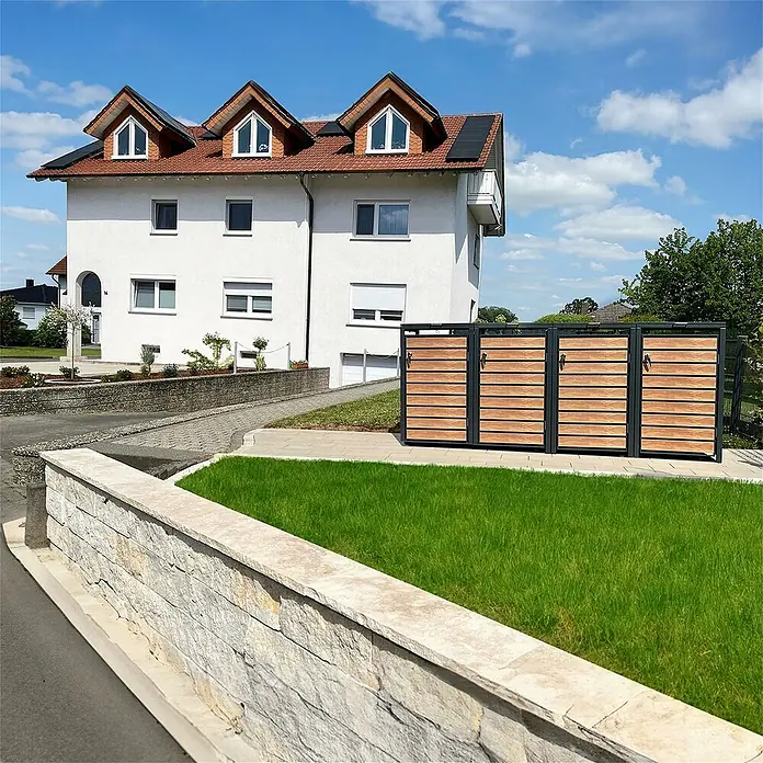 Metzler Mülltonnenbox - 272 x 80 cm, Stahl, Anthrazit/Braun, Geeignet für Anzahl Mülltonnen: 4 Stk. - Mülltonnenbox mit Holzverkleidung vor einem Haus mit rotem Dach und blauen Himmel.
