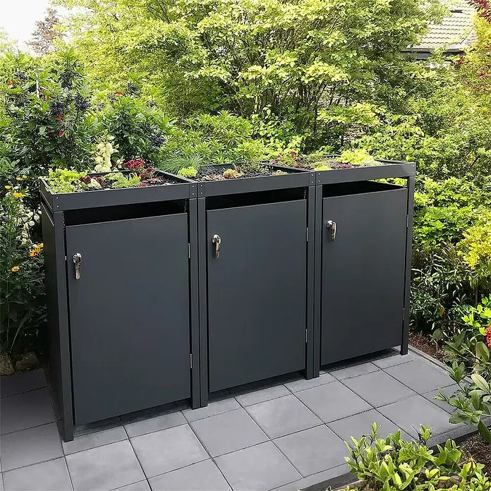 Metzler Mülltonnenbox - 198,5 x 80 cm, Stahl, Anthrazit, Geeignet für Anzahl Mülltonnen: 3 Stk., Mit Pflanzdach - Drei graue Mülltonnenboxen mit Pflanzen auf den Dächern stehen auf einem gepflasterten Bereich, umgeben von üppigem Grün.