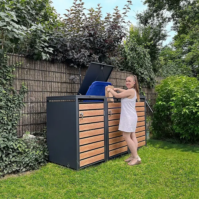 Metzler Mülltonnenbox - 204 x 80 cm, Stahl, Anthrazit/Braun, Geeignet für Anzahl Mülltonnen: 3 Stk. - Eine Frau wirft Müll in eine moderne graue Mülltonnenbox mit Holzverkleidung in einem grünen Garten. Praktische und stilvolle Aufbewahrungslösung!
