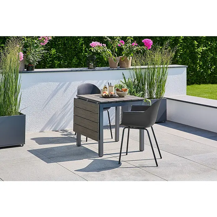 Sunfun Maja Gartentisch - L x B x H: 75 x 74,5 x 75 cm, Aluminium, Polywood, Night-Grey, Ausziehbar - Ein Gartentisch-Set mit zwei grauen Stühlen und einem quadratischen Tisch auf einer Terrasse, umgeben von Grünpflanzen.