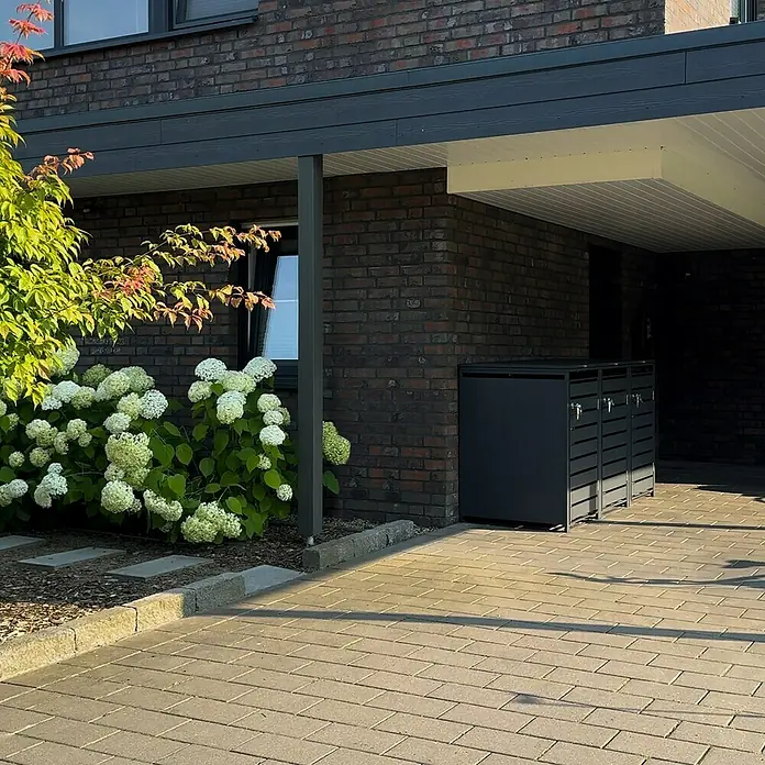 Metzler Mülltonnenbox - 204 x 80 cm, Stahl, Anthrazit, Geeignet für Anzahl Mülltonnen: 3 Stk., Mit Lamellen - Drei graue Mülltonnenboxen aus Metall stehen unter einem Carport neben einem Haus.