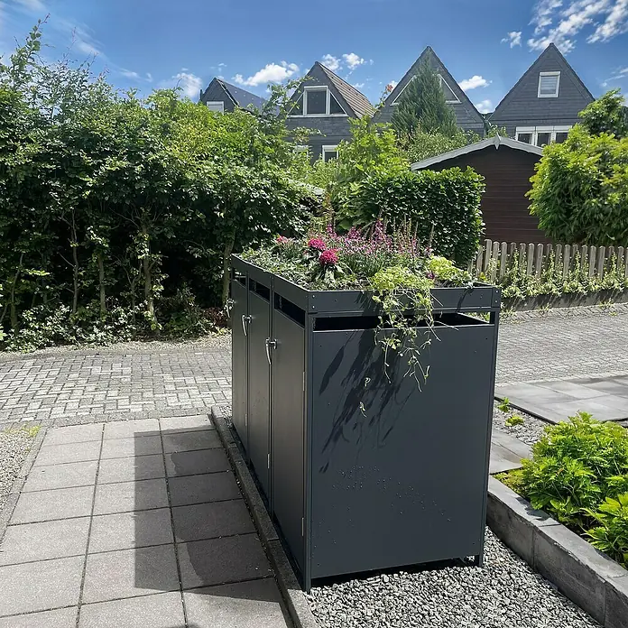 Metzler Mülltonnenbox - 198,5 x 80 cm, Stahl, Anthrazit, Geeignet für Anzahl Mülltonnen: 3 Stk., Mit Pflanzdach - Das Bild zeigt einen grauen Mülltonnenbox mit Pflanzdach, integriert in eine grüne Umgebung mit Häusern im Hintergrund.