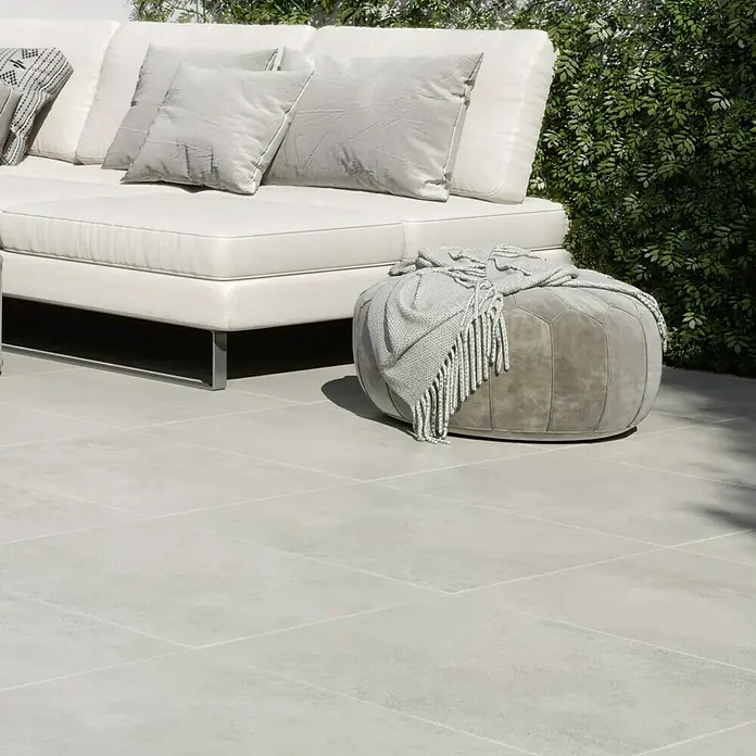 Queens Terrassenfliese - 59,3 x 119,3 x 2 cm, Weiß, Matt - Nahaufnahme eines stilvollen, modernen weißen Sofas mit grauen Kissen und einem passenden Pouf mit Decke auf einer Terrasse.