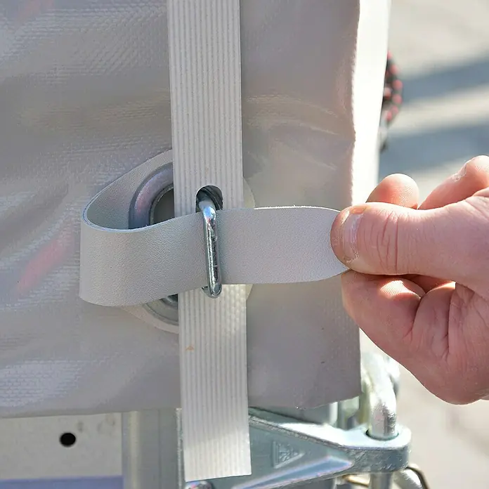 Stema Hochplanen-Set 25-13 - Passend für: Stema Anhänger Basic -25-13er-Serie, Höhe: 100 cm - Nahaufnahme einer Hand, die einen weißen Gurt mit einem Metalleisen an einer Plane befestigt.