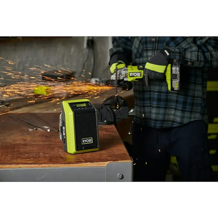 Ryobi ONE+ Akku-Bluetooth-Radio RRDAB18-0 - 10 W, Ohne Akku - Ein Ryobi-Radio steht auf einer Werkbank, während im Hintergrund jemand mit einem Winkelschleifer arbeitet und Funken sprühen.