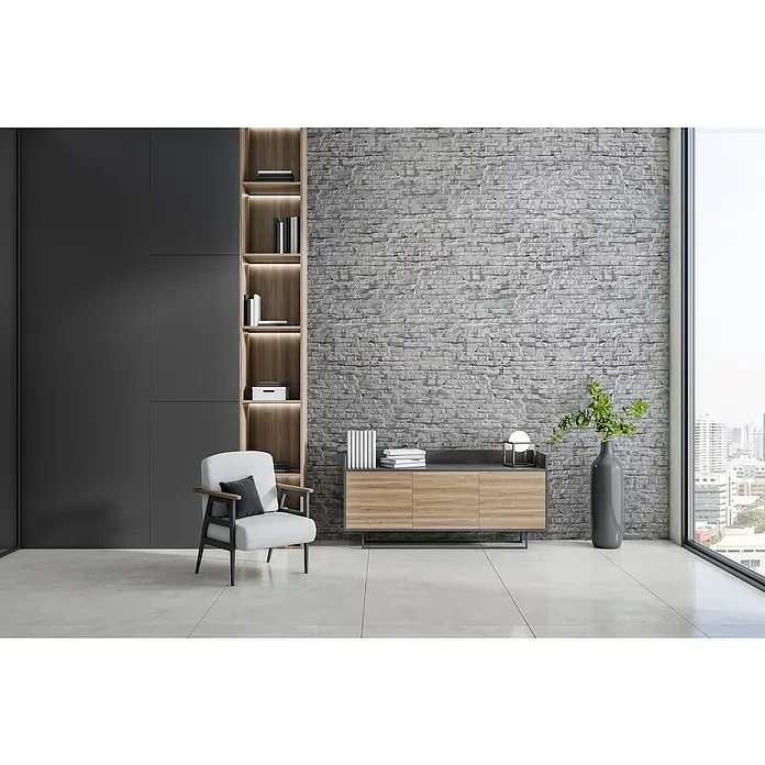 Stegu Verblendstein Hampton - 13 x 38,5 cm, Hellgrau, Steinoptik - Ein modernes Wohnzimmer mit grauem Sessel, Holzregal und stilvoller Kommode vor einer grauen Steinwand. Helles, minimalistisches Design.