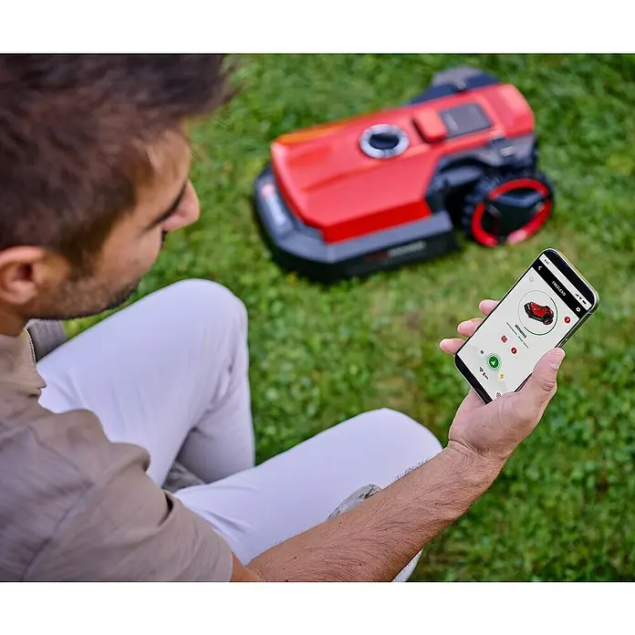 Einhell Power X-Change 18V Mähroboter Freelexo 500 - 500 m², Schnittbreite: 18 cm, Induktionsgestützte Navigation, Begrenzungskabel - Ein Mann steuert einen roten Mähroboter mit einer Smartphone-App im Grünen. Perfekt für die moderne Gartenpflege!
