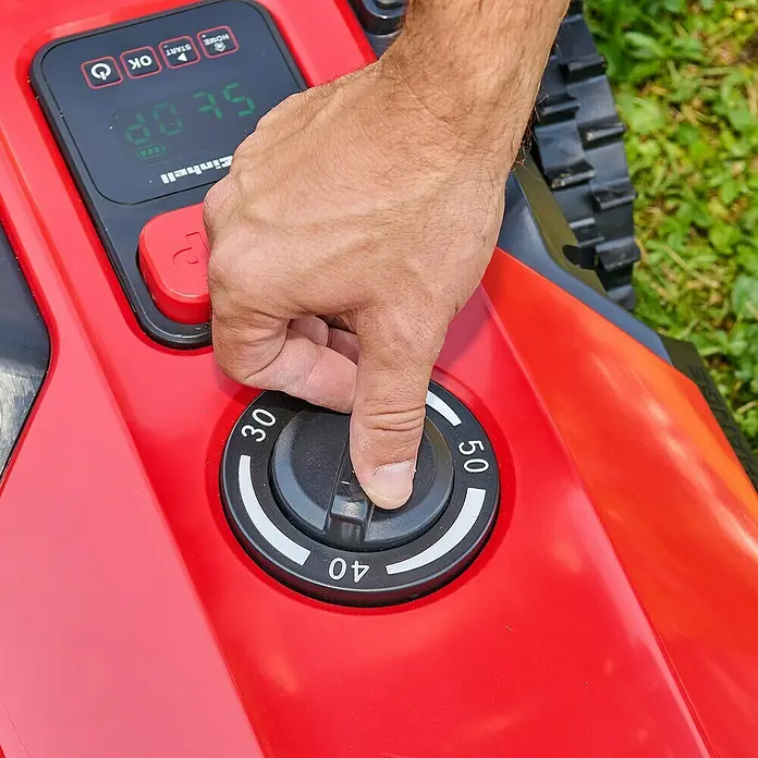 Einhell Power X-Change 18V Mähroboter Freelexo 500 - 500 m², Schnittbreite: 18 cm, Induktionsgestützte Navigation, Begrenzungskabel - Eine Hand justiert den Drehknopf eines roten Einhell Rasenmähroboters, um die Schnitthöhe einzustellen.