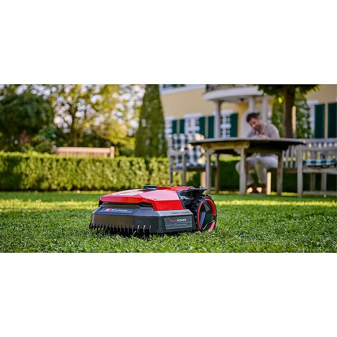 Einhell Power X-Change 18V Mähroboter Freelexo 500 - 500 m², Schnittbreite: 18 cm, Induktionsgestützte Navigation, Begrenzungskabel - Ein roter Einhell-Mähroboter steht auf einem Rasen, im Hintergrund sitzt eine Person an einem Tisch.