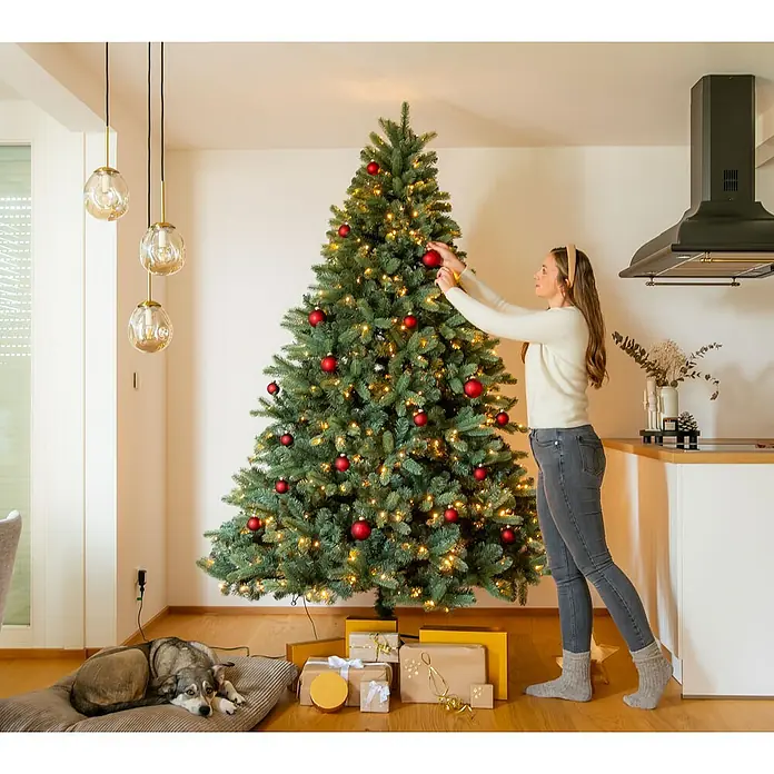 Evergreen Künstlicher Weihnachtsbaum Vermont Fichte - Höhe: 210 cm, Grün, LED-Beleuchtung - Frau schmückt einen Weihnachtsbaum, unter dem Geschenke liegen, während ein Hund daneben auf einem Kissen liegt.