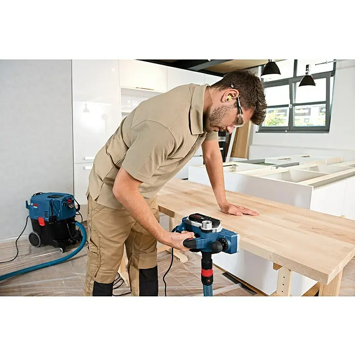 Bosch Professional Handhobel GHO 26-82 D - 710 W, Hobeltiefe: 0 mm - 2,6 mm, Leerlaufdrehzahl: 18.000 U/min - Ein Handwerker verwendet einen Bosch Elektrohobel, um eine Holzplatte zu bearbeiten. Im Hintergrund steht ein Staubsauger.