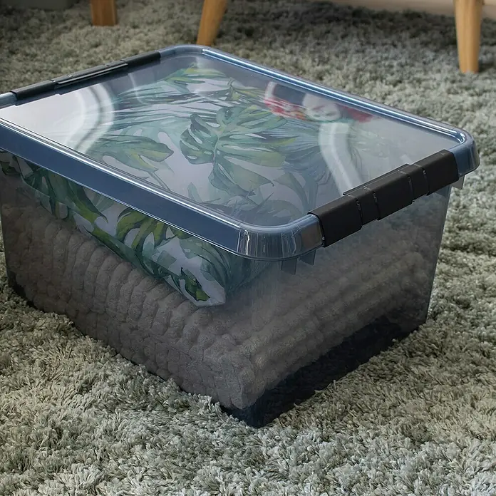 Sunware Aufbewahrungsbox Comfort Line - L x B x H: 50 x 40 x 26 cm, Transparent grau, Kunststoff - Eine durchsichtige Aufbewahrungsbox mit Deckel, gefüllt mit einer grauen Decke und einem Kissen mit Pflanzenmuster, steht auf einem grauen Teppich.