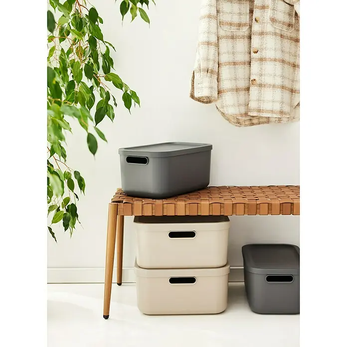 Rotho Aufbewahrungsbox - L x B x H: 39,5 x 35,5 x 17,7 cm, Kunststoff, Anthrazit, 20 l - Ein stilvolles Aufbewahrungssystem mit grauen und beigen Boxen auf einem geflochtenen Hocker und einer Jacke im Hintergrund.