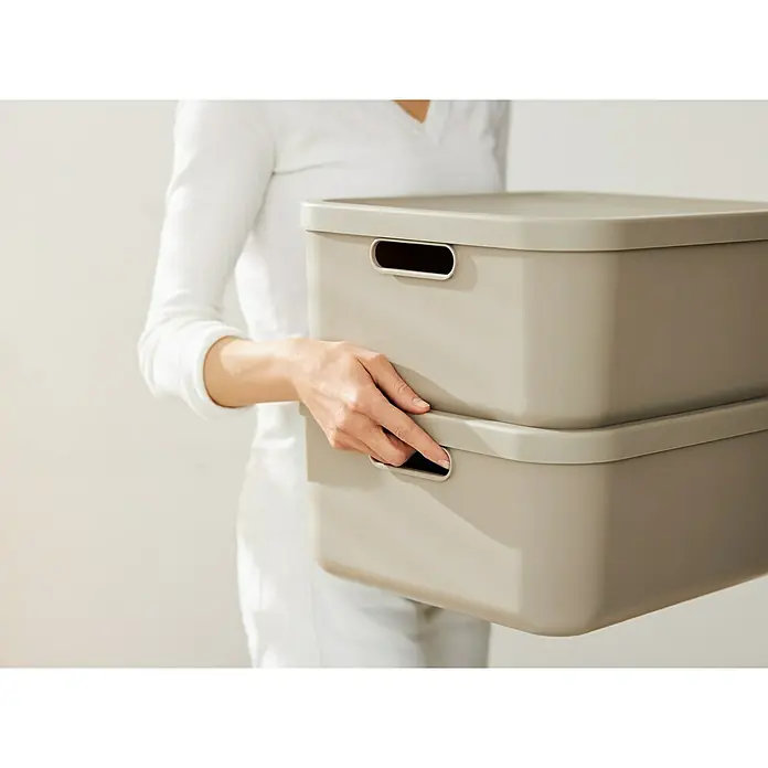 Rotho Aufbewahrungsbox - L x B x H: 39,2 x 35,5 x 17,7 cm, Kunststoff, Cappuccino, 20 l - Frau hält zwei stapelbare Aufbewahrungsboxen in Beige mit praktischen Griffen, ideal für die Organisation von Alltagsgegenständen.