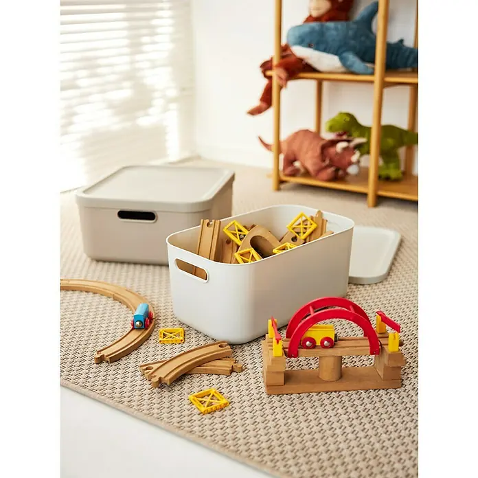 Rotho Aufbewahrungsbox - L x B x H: 39,2 x 35,5 x 17,7 cm, Kunststoff, Cappuccino, 20 l - Eine Nahaufnahme von zwei Spielzeugkisten und einer Holzeisenbahn auf einem Teppich, mit Plüschtieren im Hintergrund.