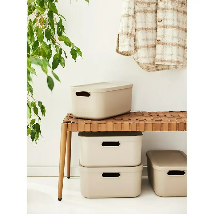 Rotho Aufbewahrungsbox - L x B x H: 39,5 x 23,5 x 17,7 cm, Kunststoff, Cappuccino, 12 l - Drei beige Kunststoffboxen mit Deckel stehen auf einem geflochtenen Hocker, eine Jacke hängt daneben. Die Boxen bieten praktischen Stauraum.