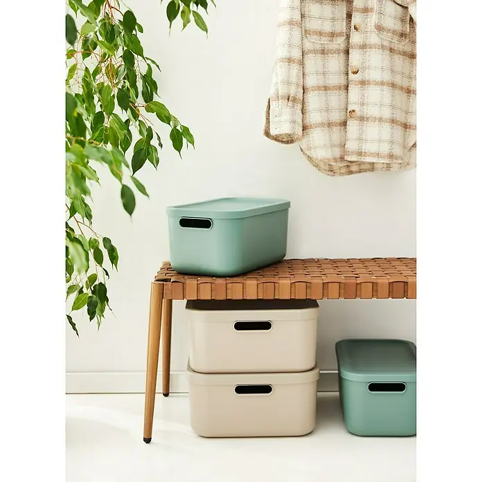Rotho Aufbewahrungsbox - L x B x H: 39,2 x 23,5 x 17,7 cm, Kunststoff, Mistelzweiggrün, 12 l - Aufnahme von drei Plastikaufbewahrungsboxen in Beige und Grün auf einem geflochtenen Ledersitzhocker vor einer weißen Wand.
