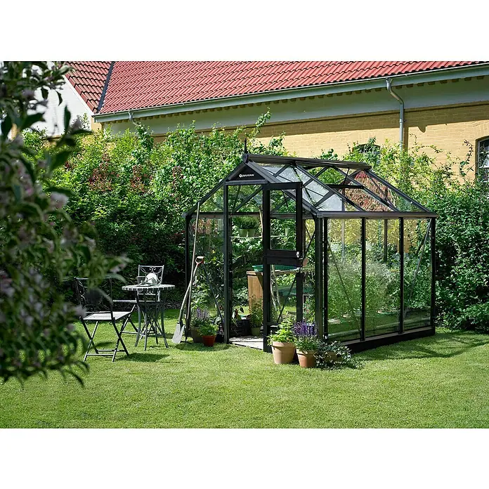 Juliana Gewächshaus Compact - Außenmaß inkl. Dachüberstand (B x T): 224 x 224 cm, Material Seitenwand: Glas, 3 mm, Schwarz - Ein Gewächshaus aus Glas steht im Garten, umgeben von Grün. Es beherbergt Pflanzen und Gartengeräte und lädt zum Verweilen ein.