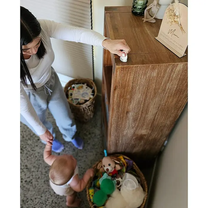 Dreambaby Magnetverschluss - Weiß, 2 Stk. - Eine Frau sichert ein braunes Holzregal mit einem weißen Schrankschloss, während ein Baby in der Nähe steht.