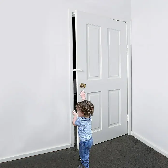 Dreambaby Fingerschutz - Weiß, 1 Stk. - Kleinkind versucht, eine weiße Tür mit goldenem Knauf zu öffnen. Erkundung und Neugier im kindlichen Alltag.