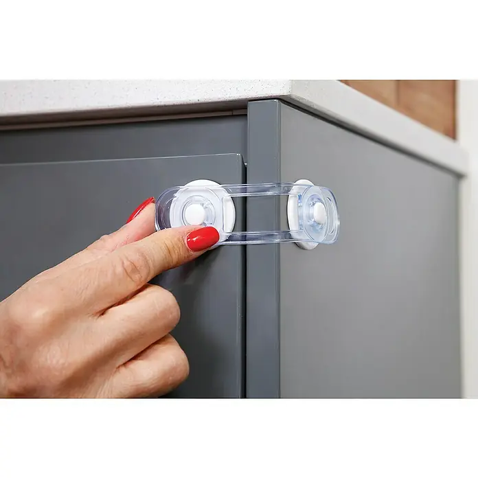 Dreambaby Mini-Multifunktionsverschluss - Kleben, Farblos, 4 Stk. - Nahaufnahme einer Hand mit roten Nägeln, die ein transparentes Schranksicherheitsschloss an einem grauen Schrank betätigt.
