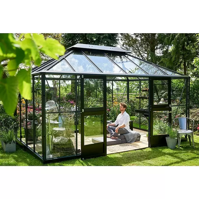 Juliana Gewächshaus Grand Oase - Außenmaß inkl. Dachüberstand (B x T): 439 x 296 cm - Schwarz gerahmtes Glasgewächshaus im Garten mit einem Mann, der darin entspannt, als persönliche Oase.