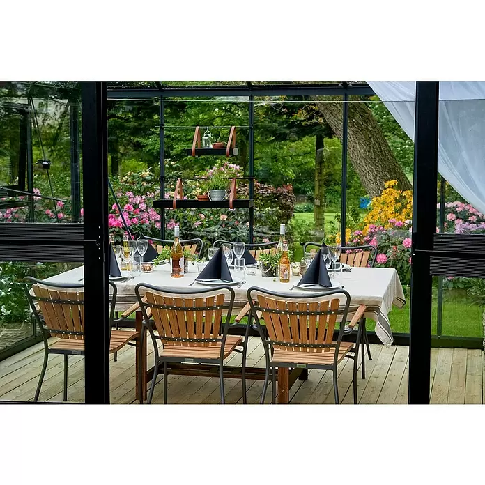 Juliana Gewächshaus Grand Oase - Außenmaß inkl. Dachüberstand (B x T): 439 x 296 cm, Material Seitenwand: Glas, 3 mm, Anthrazit - Gartensitzgruppe mit dunklen Metall- und Holzlattenstühlen um einen gedeckten Tisch in einem Glaswintergarten.