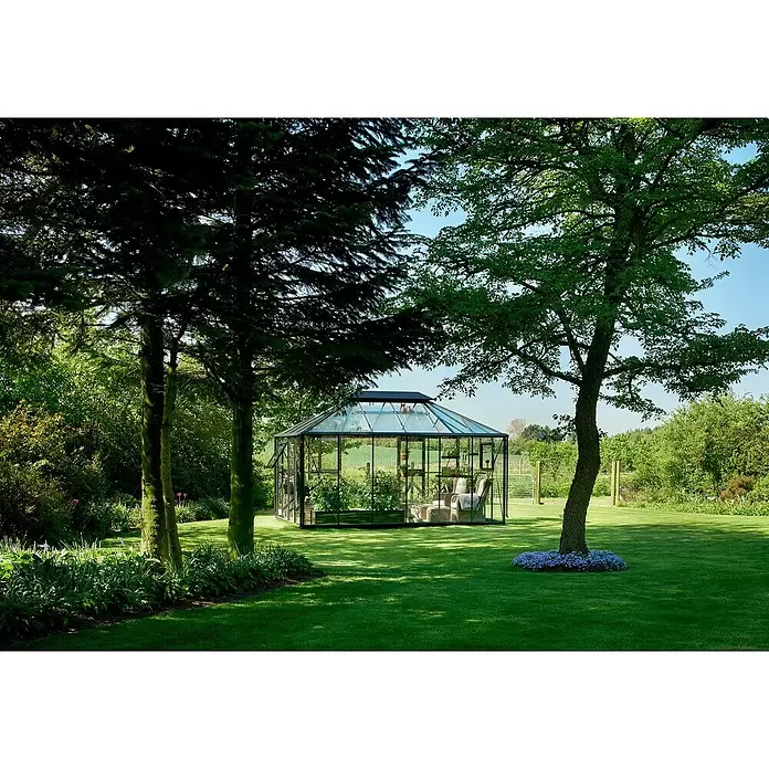 Juliana Gewächshaus Grand Oase - Außenmaß inkl. Dachüberstand (B x T): 439 x 296 cm - Gläsernes Gewächshaus mit schwarzem Rahmen in einem grünen Garten, mit Sitzbereich und Topfpflanzen.
