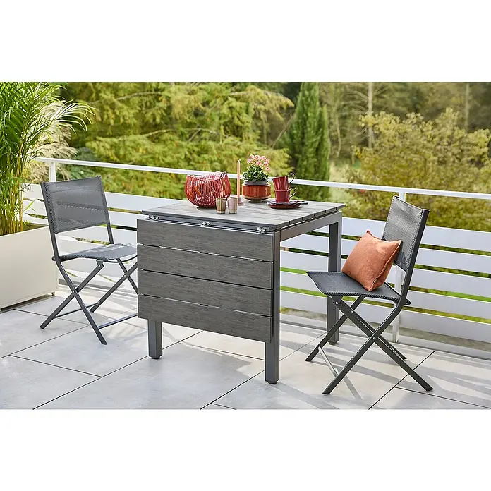 Sunfun Maja Gartentisch - L x B x H: 75 x 74,5 x 75 cm, Aluminium, Polywood, Night-Grey, Ausziehbar - Das Bild zeigt einen grauen Gartentisch mit zwei Klappstühlen auf einem Balkon. Der Tisch ist mit Dekoartikeln wie Kerzen und Pflanzen geschmückt.