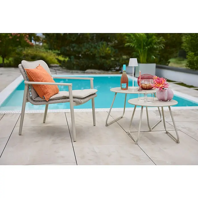 Sunfun Sarah Garten-Beistelltisch rund - Ø x H: 60 x 41,5 cm, Stahl, Light-Greige - Ein grauer Outdoor-Sessel mit orangefarbenem Kissen und zwei Beistelltischen mit dekorativen Elementen neben einem Pool.
