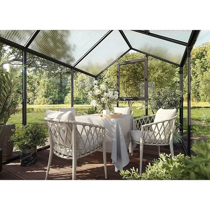 KGT Gewächshaus Aptasia IV - Außenmaß inkl. Dachüberstand (B x T): 233 x 429 cm, 4 mm, Anthrazit-Grau - Helle Gartentischgruppe mit Rattanstühlen und grauer Tischdecke in einem Gewächshaus mit Blick auf einen üppigen Garten.