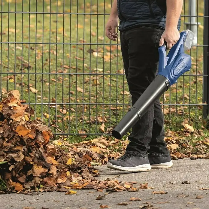 Scheppach IXES 20V Akku-Laubbläser C-BL700-X Set S - 20 V, 1 Akku, 2 Ah, Blasgeschwindigkeit: 32 m/s - 155 km/h, Blasleistung: 516 m³/h - Ein Mann benutzt einen Scheppach Laubbläser, um Blätter auf einem Gehweg zu entfernen. Das Gerät ist blau und schwarz.