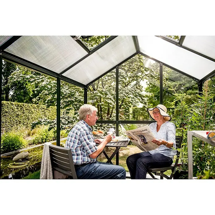 KGT Gewächshaus Galant III - Außenmaß inkl. Dachüberstand (B x T): 216 x 308 cm, Material Seitenwand: Glas, 4 mm, Tannengrün - Ein Gewächshaus mit einem Paar, das Frühstück genießt und Zeitung liest, umgeben von üppigem Grün und einem kleinen Teich.