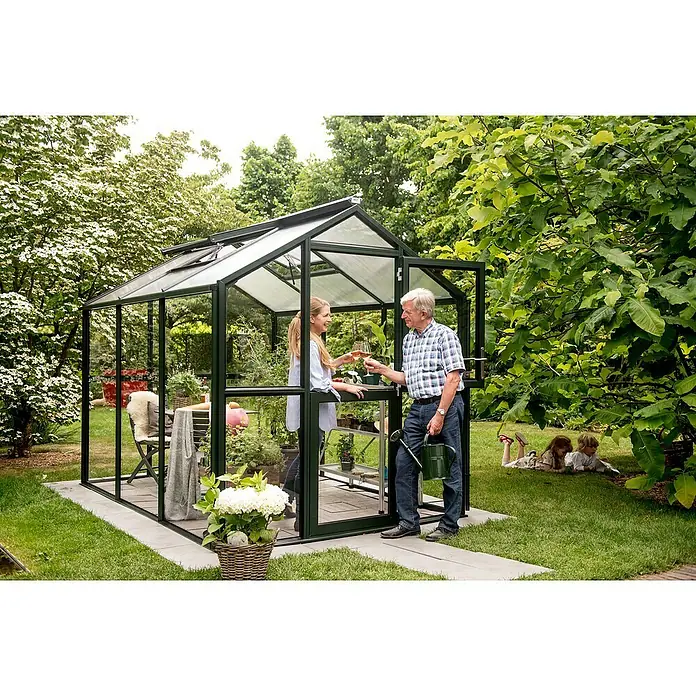 KGT Gewächshaus Galant III - Außenmaß inkl. Dachüberstand (B x T): 216 x 308 cm, Material Seitenwand: Glas, 4 mm, Tannengrün - Ein Gewächshaus mit einer Frau und einem Mann, die anstoßen, mit Kindern im Hintergrund. Das Gewächshaus bietet einen idealen Raum für Pflanzen.