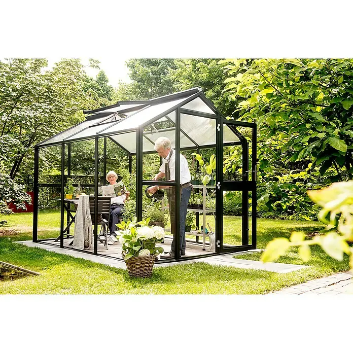 KGT Gewächshaus Galant III - Außenmaß inkl. Dachüberstand (B x T): 216 x 308 cm, Material Seitenwand: Glas, 4 mm, Tannengrün - Ein Gewächshaus aus Glas mit schwarzem Rahmen im Garten, in dem ein älteres Paar sich entspannt und die Pflanzen pflegt.